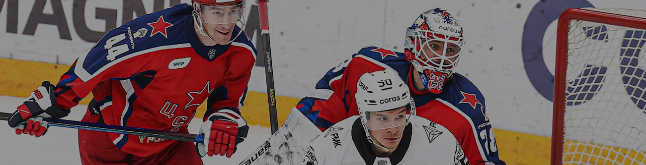 На изображении хоккеисты в момент игры: двое в красно-синей форме и один в бело-чёрной. Защитник в шлеме готовится к игре у ворот. Они сосредоточены на шайбе. На фоне видна часть рекламы на бортике