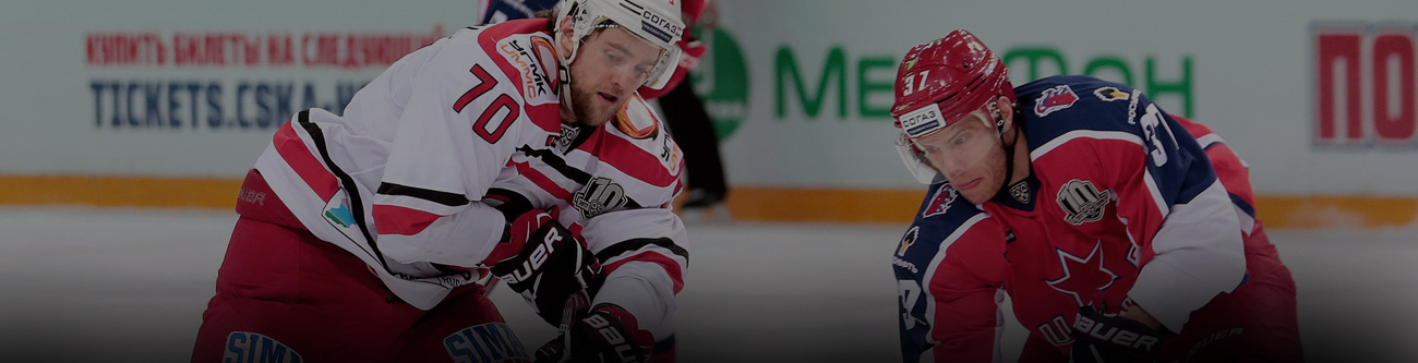 Два хоккеиста в разных формах борются за шайбу на льду. На заднем плане виден размытый рекламный баннер. Игроки сосредоточены на игре