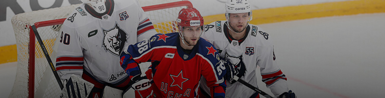 На изображении трое хоккеистов на льду. Двое в белой форме и один в красной. Они находятся рядом с воротами, натянуты сетки. Хоккеисты сосредоточены на игре. На форме видны номера и эмблемы