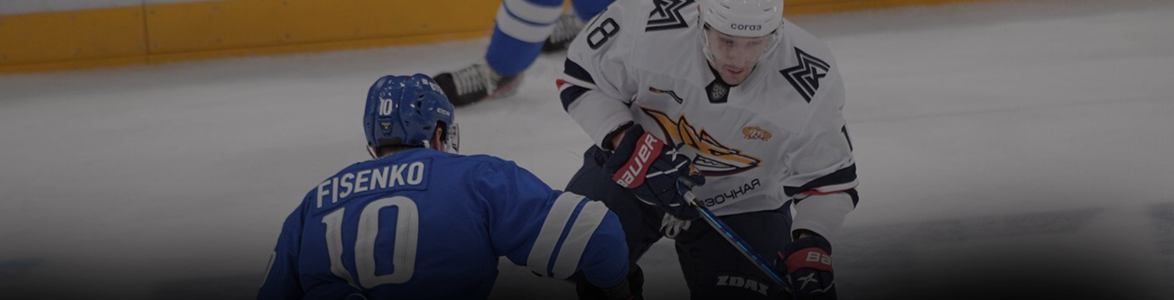 Хоккеисты на льду участвуют в матче. Игрок в синей форме с номером 10 и надписью Фисенко на спине противостоит игроку в белой форме. На льду видно хоккейную клюшку