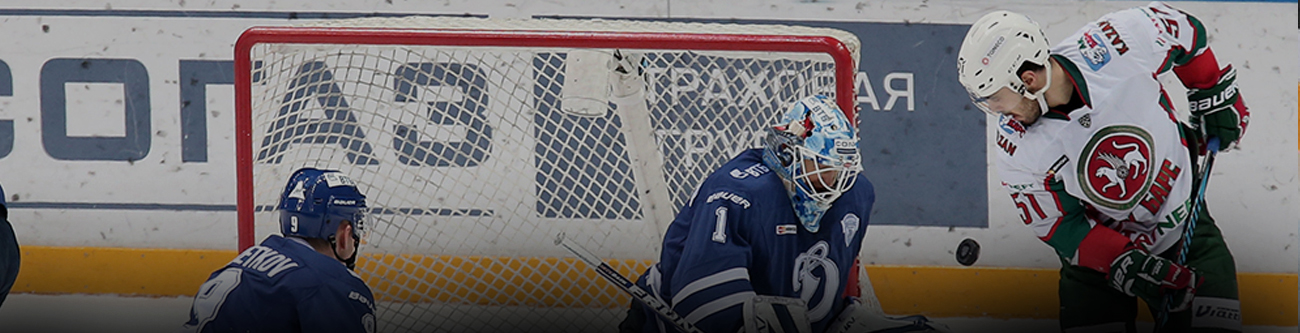 На изображении хоккейный матч: один игрок в зелёно-красной форме атакует ворота, за которыми стоит вратарь в синей форме. На фоне видно рекламу и хоккейного полевого игрока