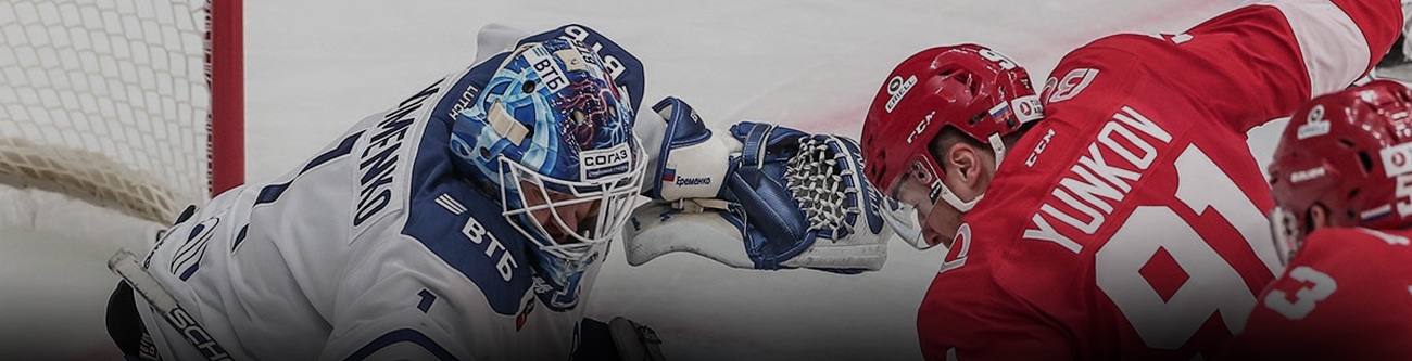 На изображении хоккейный вратарь в синей форме пытается сделать сейв, преграждая путь шайбе, пока игрок в красной форме атакует ворот. На заднем плане видны ворота