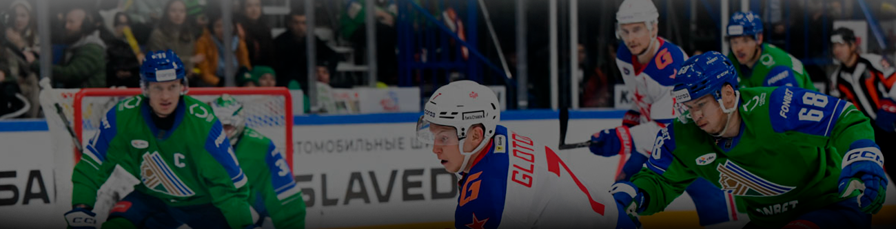 На изображении хоккейный матч. Игроки в зелёной форме активно обороняются против команды в бело-синей форме. Оживлённая атмосфера с болельщиками на трибунах. Видно напряжённый момент игры на ледовой площадке