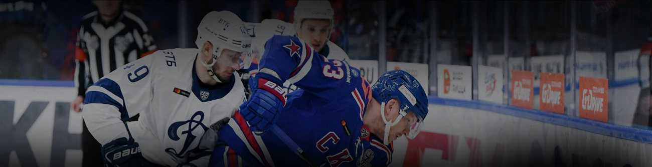 На хоккейной площадке игрок в синей форме преследует игрока в белой форме у борта. Судья наблюдает за игрой. Атмосфера напряжённой борьбы в матче