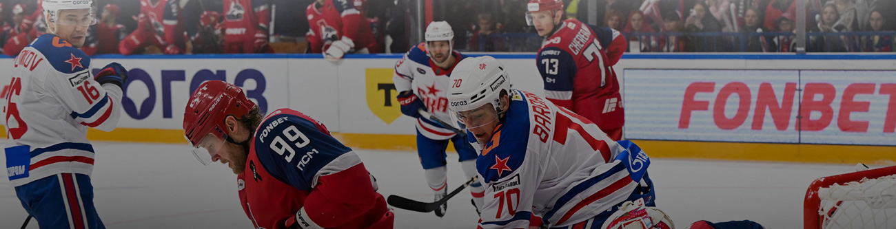 На изображении хоккейный матч, где игроки из команды в красной форме сражаются против команды в белой форме с синими и красными деталями. Один из игроков атакует, в то время как другие следят за шайбой. На заднем плане видны зрители