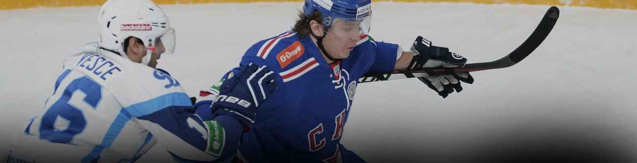 На изображении запечатлен момент хоккейного матча: игрок в синей форме с клюшкой противостоит сопернику в бело-голубой форме. Активная борьба за шайбу на льду