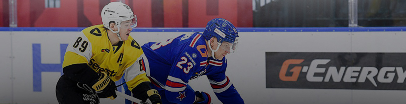 На изображении два хоккеиста в динамичной борьбе за шайбу на льду. Игрок в жёлтой форме рядом с игроком в синей форме. На заднем плане видны бортики с рекламными баннерами