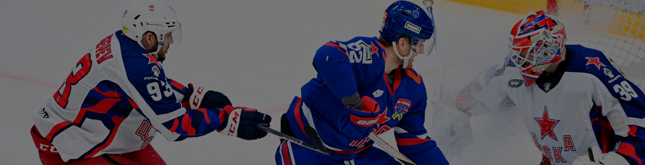 На изображении запечатлен хоккейный матч. Три игрока на льду: двое соперников в разных формах и вратарь в шлеме защищают ворота. Игра идет в динамике, команда в синей форме атакует