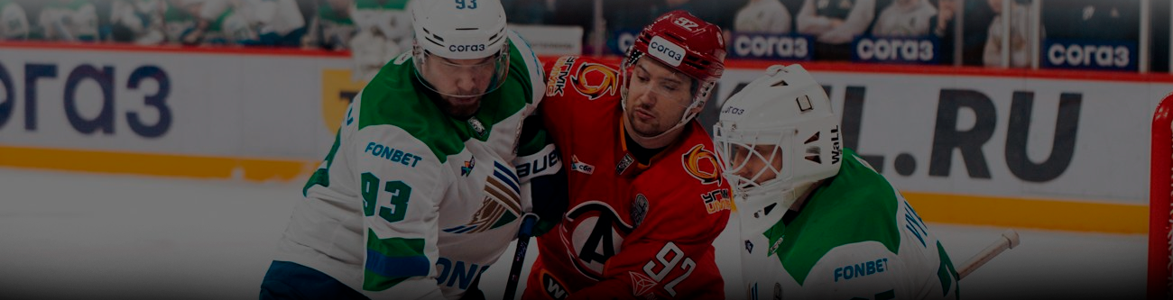 Хоккейный матч: игрок в красной форме борется за шайбу с двумя игроками в бело-зеленой форме. На заднем плане видна рекламная вывеска и зрители на трибуне