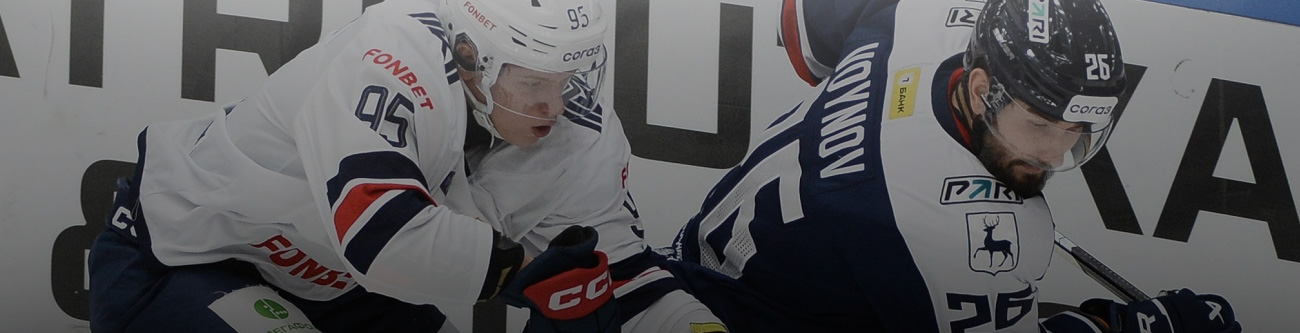 На изображении два хоккеиста в игровой форме борются за шайбу у борта на льду. Слева спортсмен в белой форме, справа — в темной. На заднем плане часть рекламного баннера