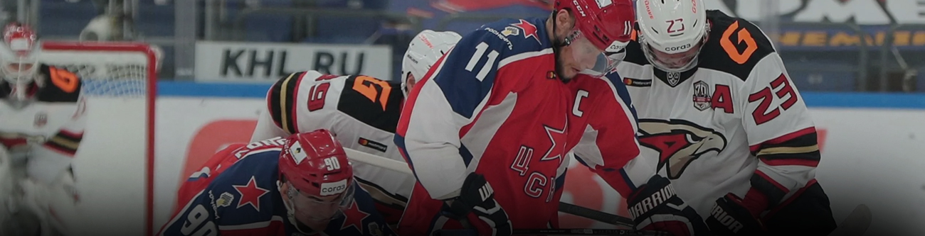 На изображении запечатлен момент хоккейного матча. Трое игроков борются за шайбу на льду. Спортсмены одеты в разные формы: красно-синюю и бело-красно-черную. На заднем плане видна надпись KHL