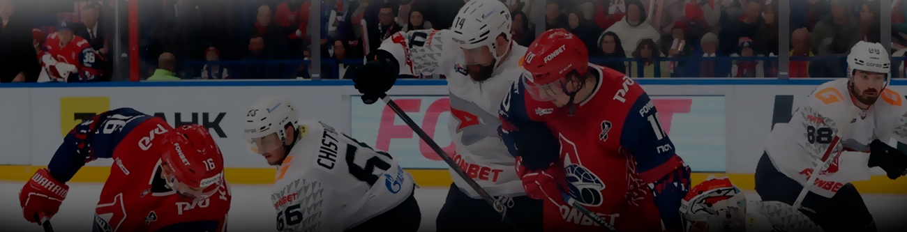 На изображении запечатлен хоккейный матч: игроки в форме красного и белого цветов активно борются за шайбу на льду. На заднем плане видны зрители на трибунах