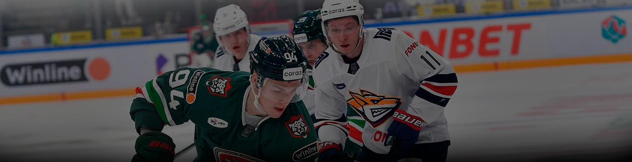 На изображении хоккейный матч: игроки команды в зелёной форме против игроков в белой форме с элементами синего и оранжевого. Видна часть ледовой арены и рекламные баннеры на заднем плане