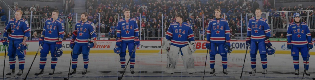 Хоккейная команда в синей форме стоит на льду перед началом игры. Игроки выстроились в линию с клюшками, на заднем плане видны трибуны с болельщиками